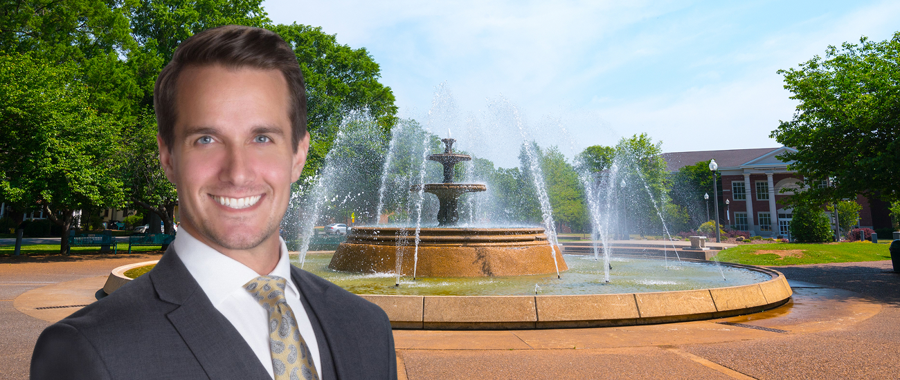 Smiling attorney in a suit pictured in front of a large fountain and office building.