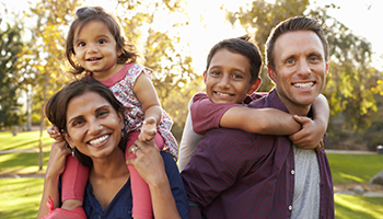 Smiling parents carry their two young children on their backs in a sunny park.