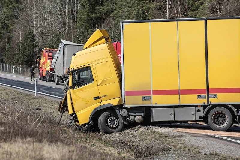 Damaged yellow semi-truck crashed on the side of a highway with tow trucks in the background